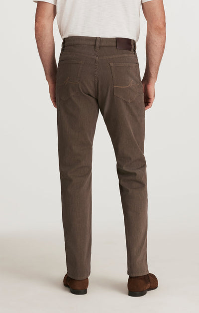A man is seen from behind wearing 34 Heritage Courage Diagonal Pants in a coffee color, paired with a white shirt and brown shoes. The focus is on the straight fit and distinctive diagonal weave of these 34 Heritage pants.