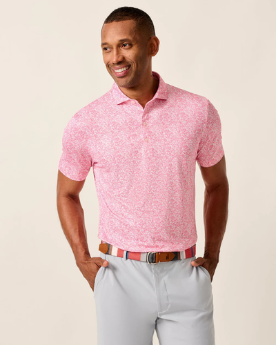 A man wears the Johnnie-O Performance Polo- Wildflower by Johnnie-O, featuring a light pink floral pattern and UPF 50 sun protection, paired with gray pants and a striped belt, standing with hands in his pockets against a beige background.