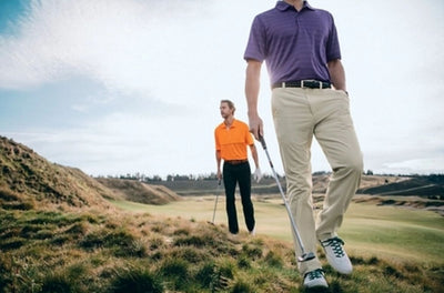 Two men with golf clubs walk on a grassy course. One wears a purple shirt and beige Cutter & Buck Bainbridge Pants Big & Tall, while the other sports an orange shirt and black flat front pants.