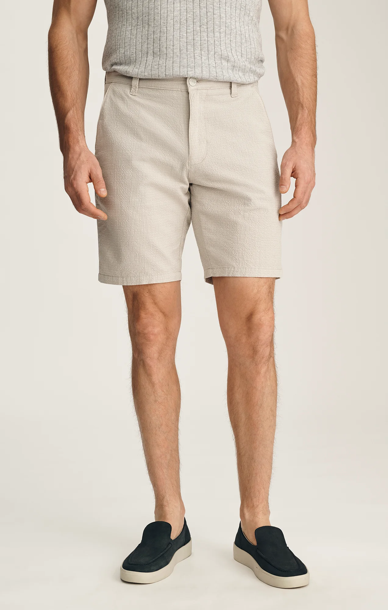 A man wears 34 Heritage Arizona Seersucker Shorts in beige, a light gray ribbed shirt, and black slip-on shoes, standing against a plain background.