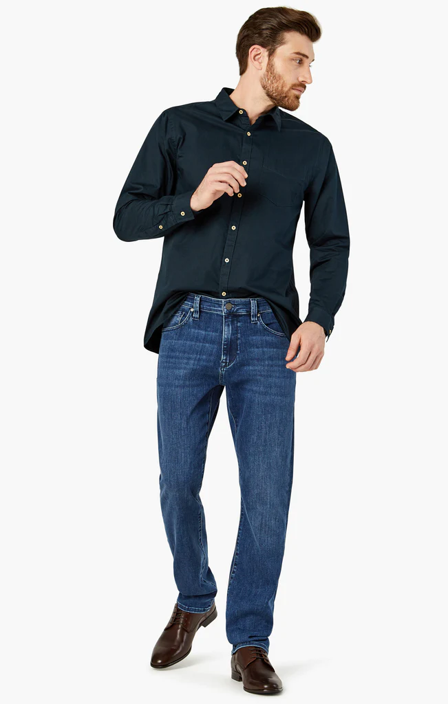 A man wearing a dark button-up shirt, 34 Heritage Charisma Mid Urban relaxed fit straight leg jeans, and brown shoes stands sideways against a plain white background.