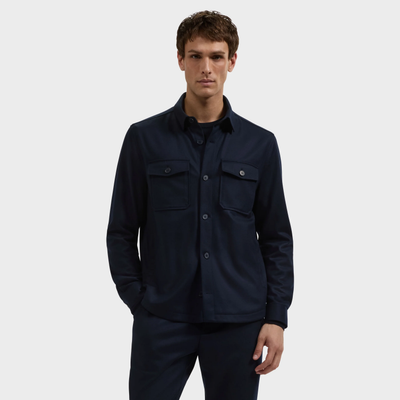 A man stands against a plain background, wearing the Bugatchi Button Shirt Jacket KX9015JS1 in navy with two front pockets, paired with matching navy pants.