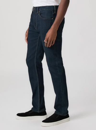 Wearing Paige Federal Straight Slim Jeans in dark blue by Paige, paired with a white shirt and black slip-on shoes, a person stands against a plain light background.