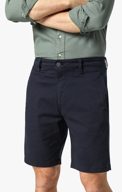 A person stands with arms crossed, wearing a green long-sleeve shirt with rolled sleeves and dark navy 34 Heritage Nevada Twill Shorts by 34 Heritage, made from breathable stretch fabric.