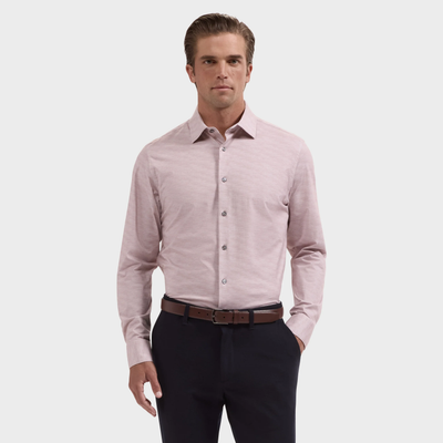 A man wearing the Bugatchi James Herringbone Sport Shirt (style KF9508K60) in light pink, paired with dark pants and a brown belt, stands with one hand in his pocket against a plain background.