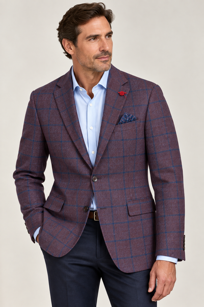 A man with brown hair and a trimmed beard stands with one hand in his pocket, looking to the side. He is wearing the TailoRed Maroon Navy Windowpane Wool Sport Coat by TailoRed, expertly crafted from fine Italian fabric, paired with a light blue shirt and navy pants.