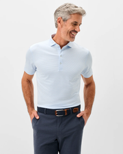 A man with gray hair smiles with his hands in his pockets, wearing the Johnnie-O Lyndonn Stripe Performance Knit Polo by Johnnie-O in light blue and dark pants, standing slightly to the side.