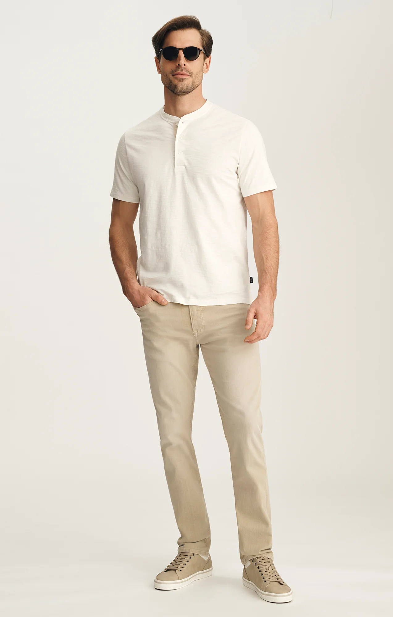 A man wearing sunglasses, a short-sleeve white Henley shirt, 34 Heritage Courage Jeans Urban by 34 Heritage, and beige sneakers stands against a plain light background.