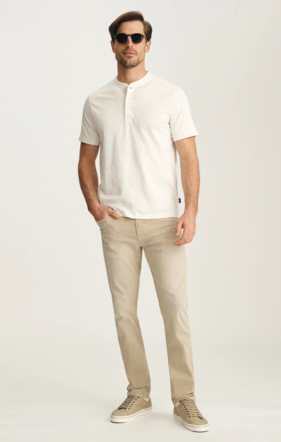 A man wearing sunglasses, a short-sleeve white Henley shirt, 34 Heritage Courage Jeans Urban by 34 Heritage, and beige sneakers stands against a plain light background.
