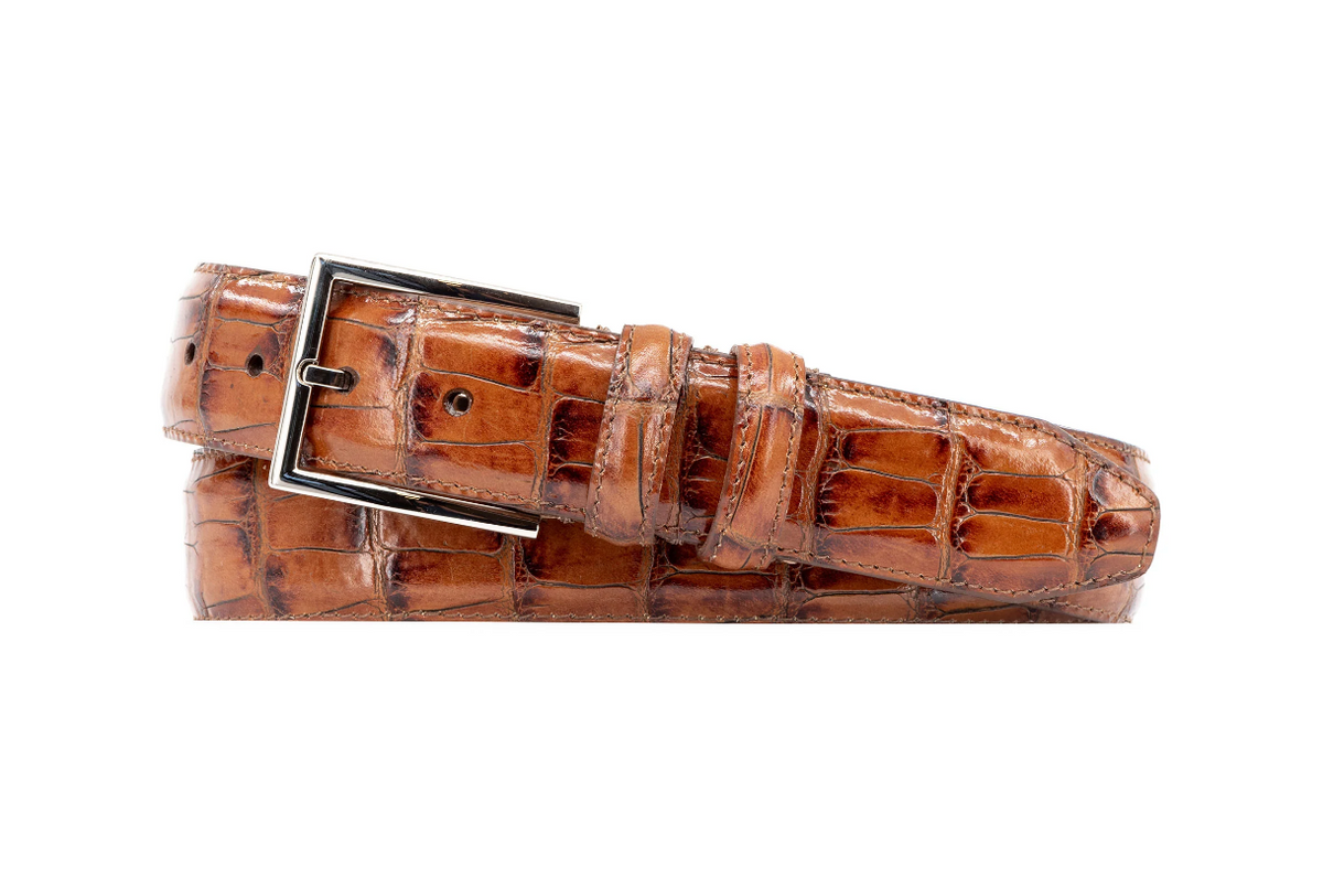 The Martin Dingman Alexander Alligator is a brown alligator grain leather belt with a silver rectangular buckle, rolled up against a white background.