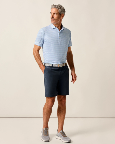 A man stands against a plain background wearing a light blue striped polo, Johnnie-O Mulligan Big & Tall Shorts in dark blue, a belt, and gray sneakers.