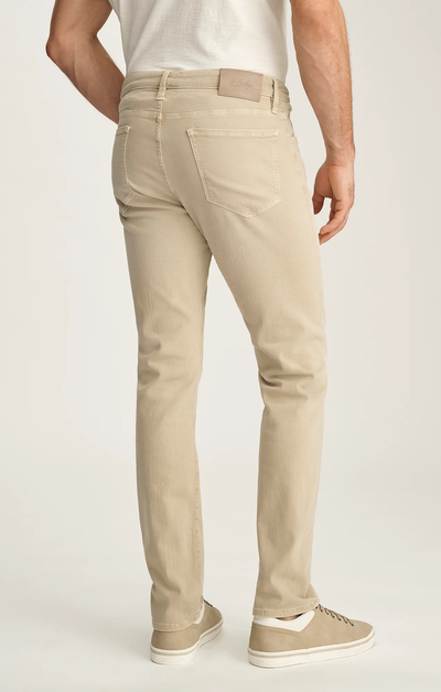 A man wearing 34 Heritage Courage Jeans Urban in beige, crafted from breathable Tencel, paired with a white t-shirt and beige sneakers, stands facing away to display the back view of the jeans by 34 Heritage.