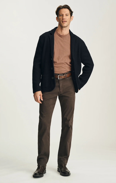 A man stands facing forward against a plain light background, wearing a black textured blazer, brown t-shirt, 34 Heritage Courage Twill Pant in brown by 34 Heritage, and dark brown shoes.