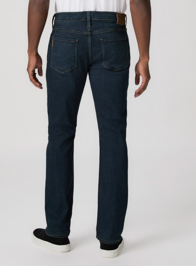 A person is seen from the back wearing Paige Federal Straight Slim Jeans by Paige in a dark wash, paired with a white shirt and black shoes against a plain background.