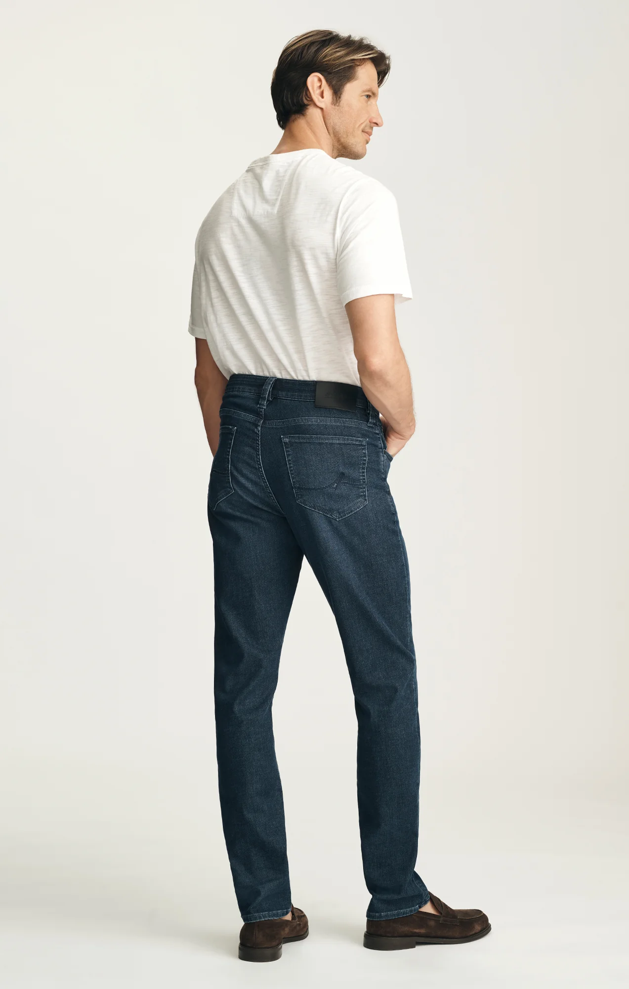 A man stands facing away, wearing a white T-shirt, brown shoes, and 34 Heritage Courage Sporty Jeans in dark blue—crafted with Thermocool fiber technology—against a plain light background.