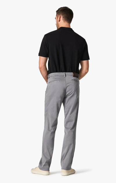 A man stands facing away, wearing a black short-sleeve polo shirt, white sneakers, and 34 Heritage Charisma Shark Twill Big & Tall light gray straight leg chino pants by 34 Heritage against a plain white background.