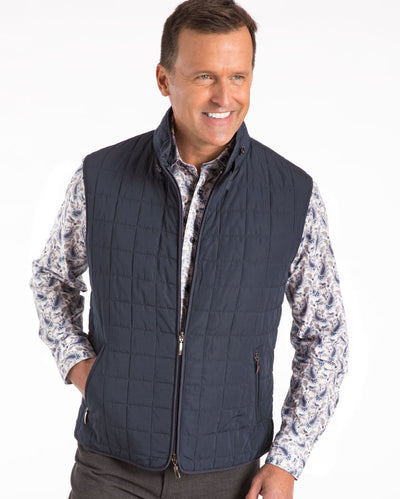 A man wears the St Croix Quilted Vest 7051 by St. Croix over a patterned button-up shirt, standing with one hand in his pocket and smiling against a plain white background.
