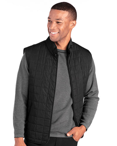 A man wearing the St. Croix Quilted Vest 7051 over a gray long-sleeve shirt stands with one hand in his pocket, looking off to the side and smiling.