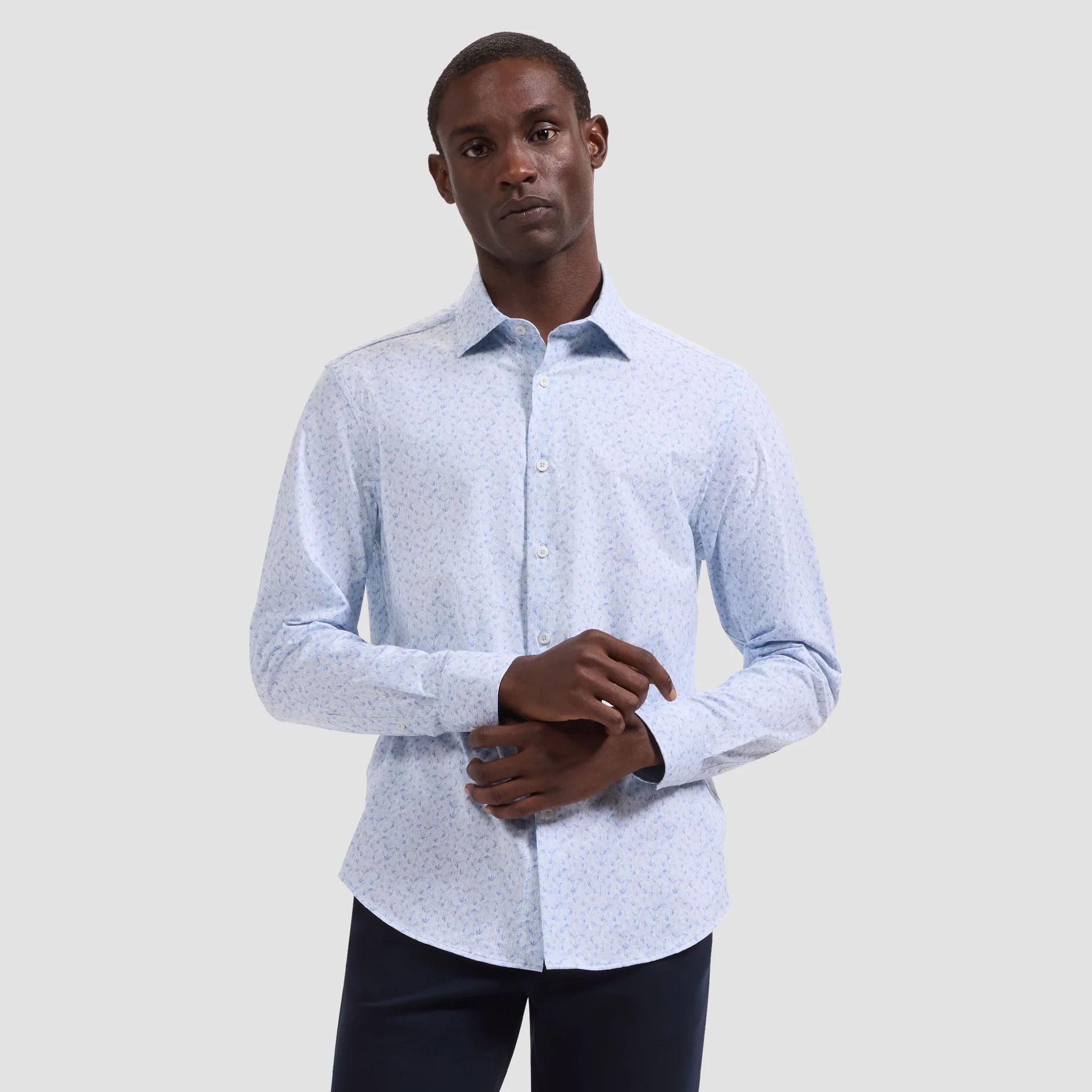 A man in a light blue patterned Bugatchi OP Jimmy OoohCotton® Shirt JF9648K61, made from wrinkle-resistant performance fabric, and dark pants stands before a plain light gray background.