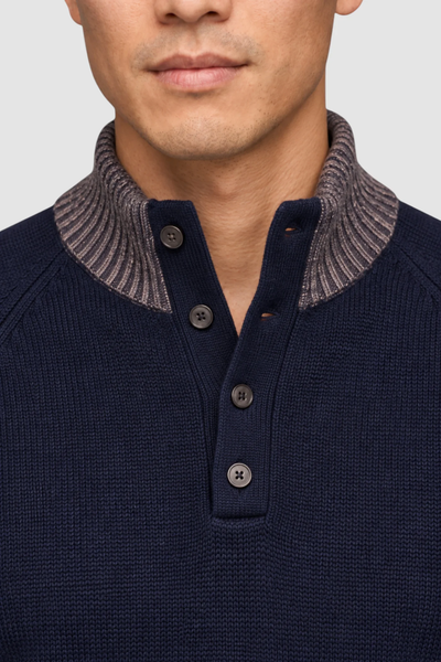 Close-up of a person wearing the Garnet Tate Contrast-Trim Mock Neck Sweater 2622022 by Garnet, a navy blue men’s wool-blend sweater with a gray ribbed collar and four black buttons.