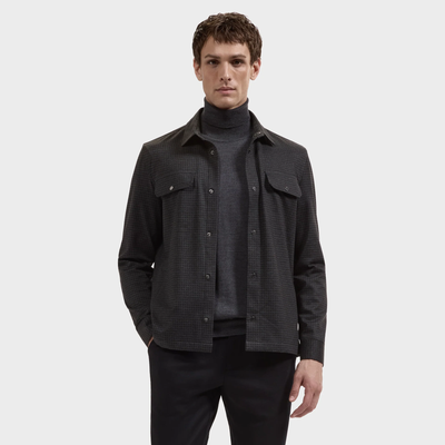 A man wearing the Bugatchi Micro Check Knit Shirt KF2313K93 layered over a charcoal long-sleeve performance stretch knit shirt, paired with black pants, stands with one hand in his pocket against a plain white background.
