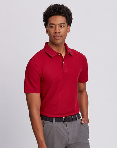 A man wears a red Cutter & Buck Breakthrough Polo Big & Tall (UPF 50+) and gray pants with a black belt, standing against a plain light background.