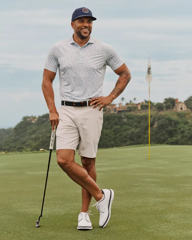 A man stands on a golf course wearing a Johnnie-O Cabana Bar Performance Jersey Polo, a cap, light shorts, and golf shoes, holding a putter with lush greenery and a flag in the background.