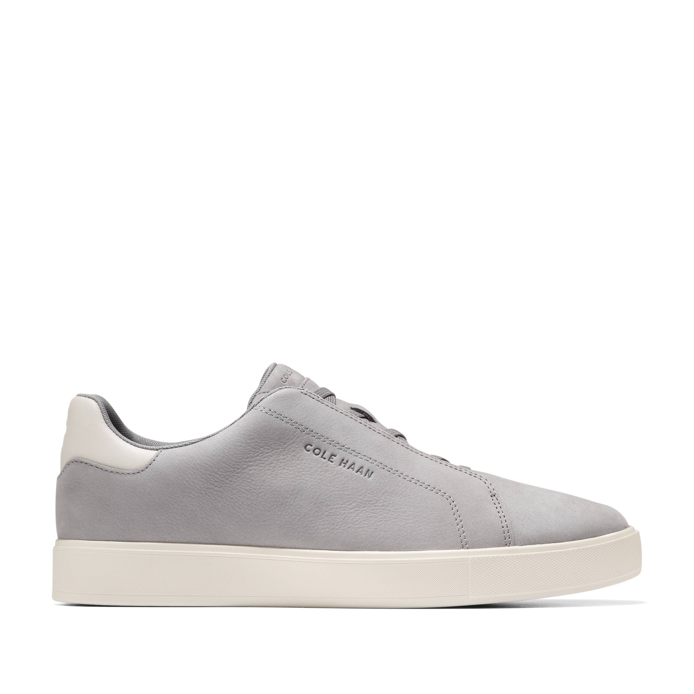 The Cole Haan GrandPro Luxe Slip On Sneaker is a light grey leather shoe with a breathable upper, white sole, and heel tab. Featuring Cole Haan branding on the side, it is shown in a side view against a plain white background.