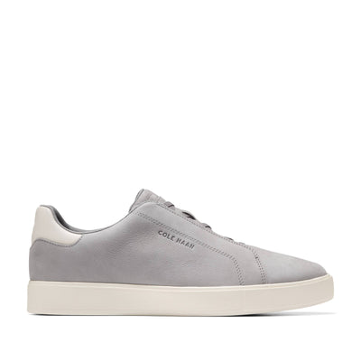 The Cole Haan GrandPro Luxe Slip On Sneaker is a light grey leather shoe with a breathable upper, white sole, and heel tab. Featuring Cole Haan branding on the side, it is shown in a side view against a plain white background.