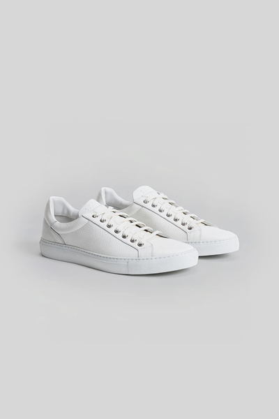 The Robert Barakett Terrence Leather Sneaker by Robert Barakett is a white low-top dress-casual shoe with white laces and a flat sole, made in Italy, shown on a light gray background.