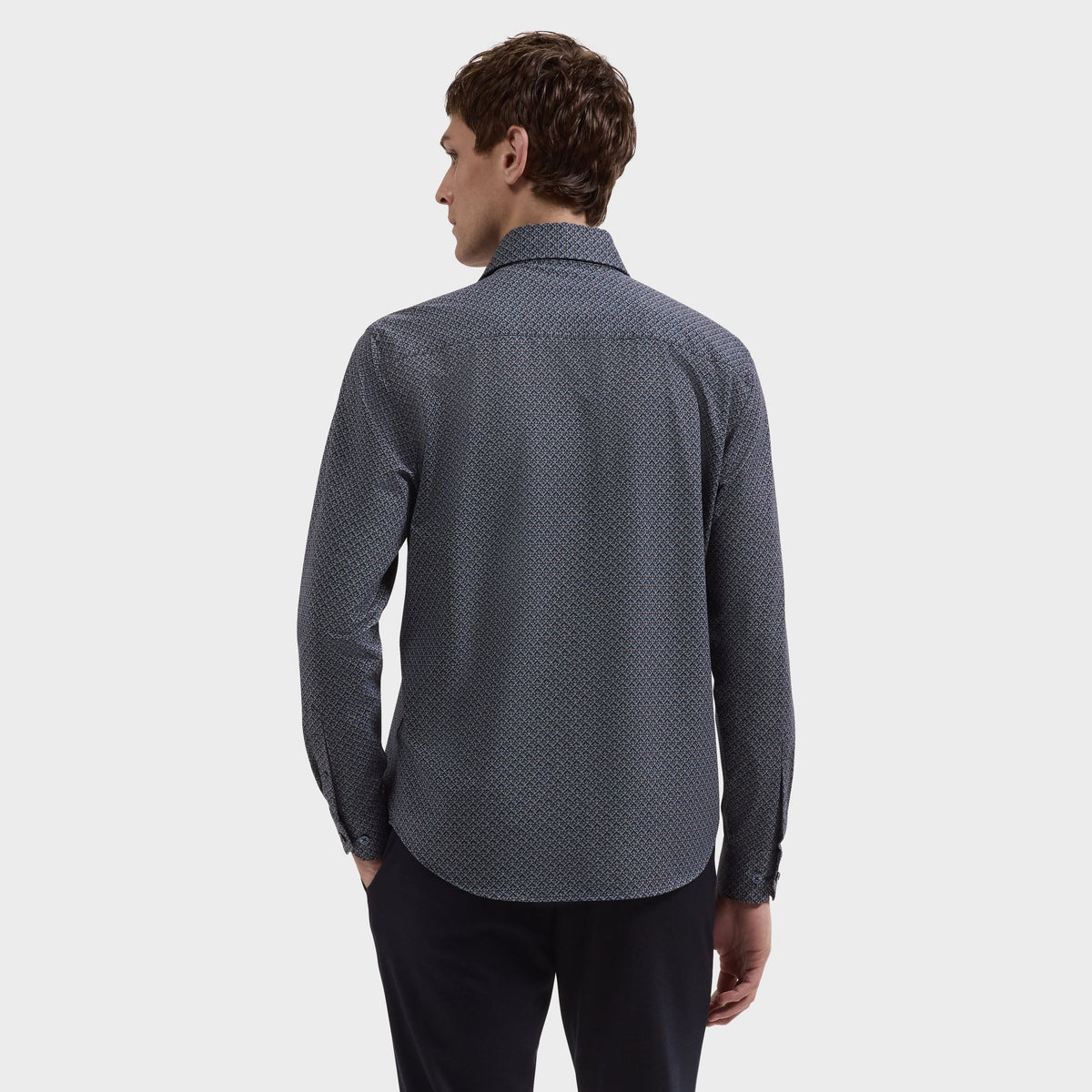 A man with short brown hair, dressed in a dark, long-sleeve Bugatchi Devon Geometric Sport Shirt (KF9431K80) and black pants, stands with his back to the camera against a plain white background.