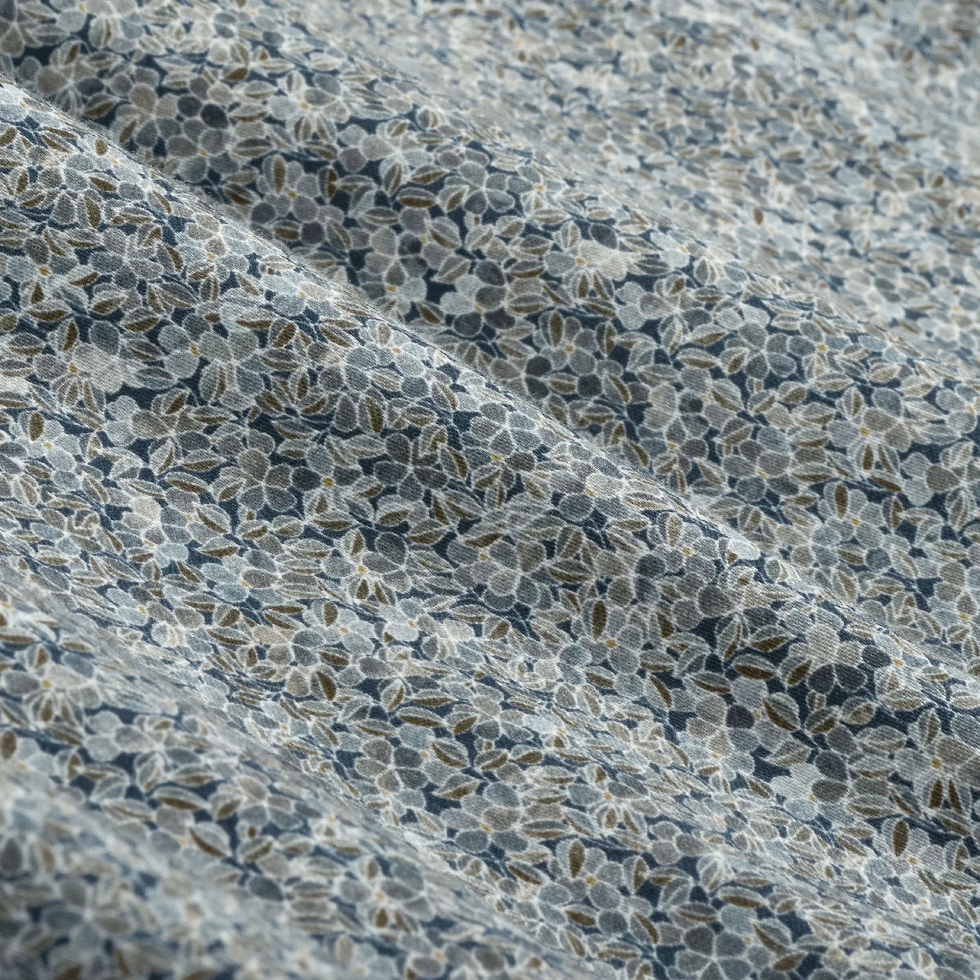 Close-up of light blue fabric adorned with a small floral pattern in shades of blue, white, and olive green, highlighting gentle folds and texture—an elegant detail featured in the Bugatchi Alvin Floral Sport Shirt KS3128L80S by Bugatchi, designed for wrinkle-resistant wear throughout the day.