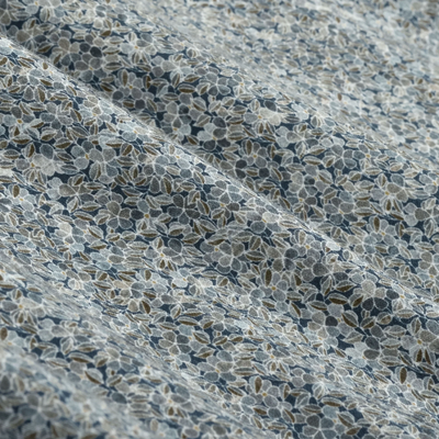 Close-up of light blue fabric adorned with a small floral pattern in shades of blue, white, and olive green, highlighting gentle folds and texture—an elegant detail featured in the Bugatchi Alvin Floral Sport Shirt KS3128L80S by Bugatchi, designed for wrinkle-resistant wear throughout the day.