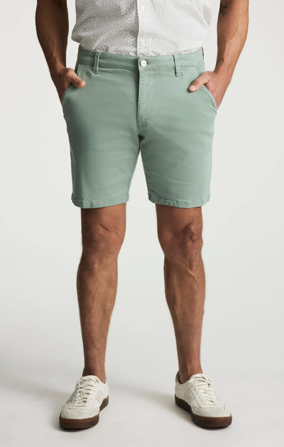 Man wearing 34 Heritage Arizona Twill Short in mint green, a white short-sleeve button-up shirt, and white sneakers, standing with hands in pockets against a plain background.