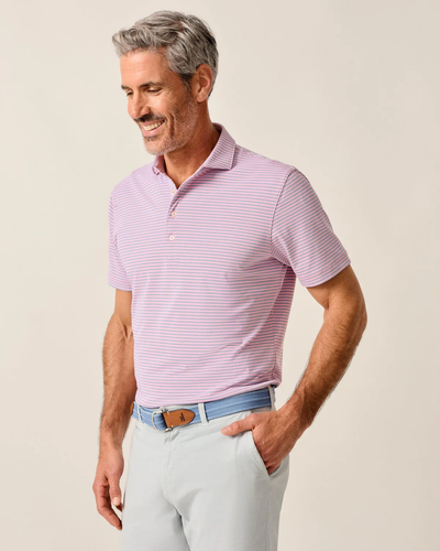 A man with gray hair and a beard wears the Johnnie-O Performance Polo-Stetsons by Johnnie-O in light pink and white stripes (UPF 50), paired with light gray pants and a blue belt, standing with one hand in his pocket.