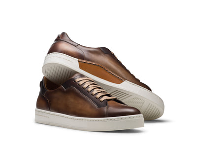 The Magnanni Amadeo Sneaker by Magnanni features brown sporty leather with white soles; one shoe stands upright while the other lies on its side, showcasing both the sole and side profile.