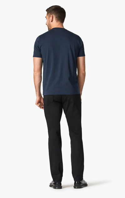 A man stands facing away from the camera, wearing a navy blue t-shirt, 34 Heritage Charisma Double Black pants by 34 Heritage with a higher-rise fit, and black shoes against a plain white background.