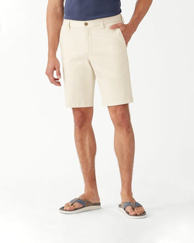 Wearing Tommy Bahama Boracay Shorts Big & Tall in beige, a person stands with one hand in their pocket, paired with a navy shirt and gray flip-flops against a white background.
