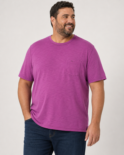 A man wearing the Tommy Bahama Bali Beach Crew Tee in bright purple, featuring short sleeves, a crew neck, and a chest pocket, paired with dark jeans and standing against a plain light background.