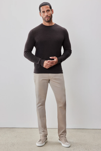 A man stands against a light-colored wall, wearing the Robert Barakett Watertown Sweater by Robert Barakett, beige pants, and light sneakers.