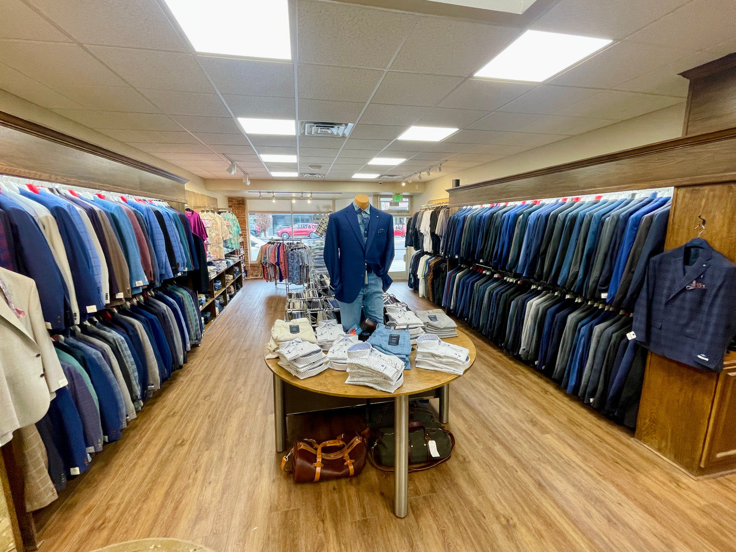 Interior of a mens clothing store with racks of suits on both sides, a display table with shirts in the center, and a mannequin wearing a blue blazer and jeans.