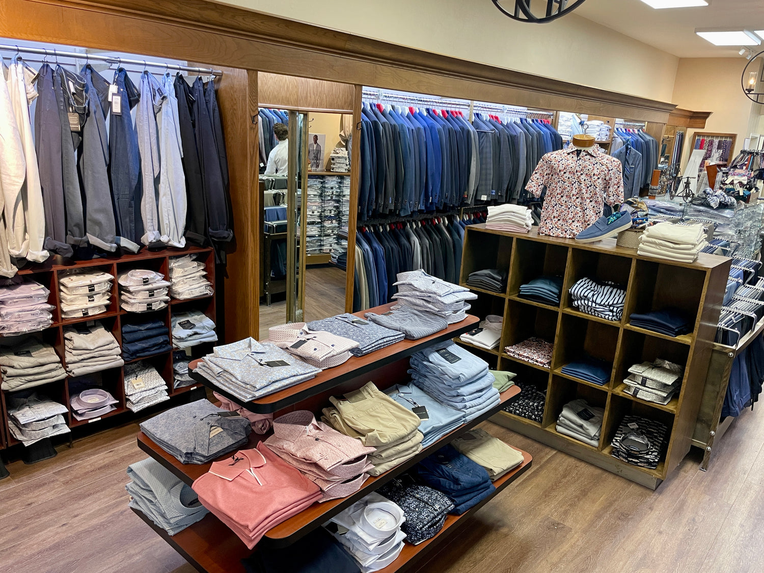 Men’s clothing store interior with folded shirts and pants on shelves, suit jackets hanging in the background, and a fitting room visible with a large mirror.