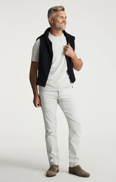A man with gray hair wearing a white t-shirt, 34 Heritage Courage Jeans Organic Ultra by 34 Heritage, brown shoes, and a black sweater draped over his shoulders stands and smiles against a plain background.