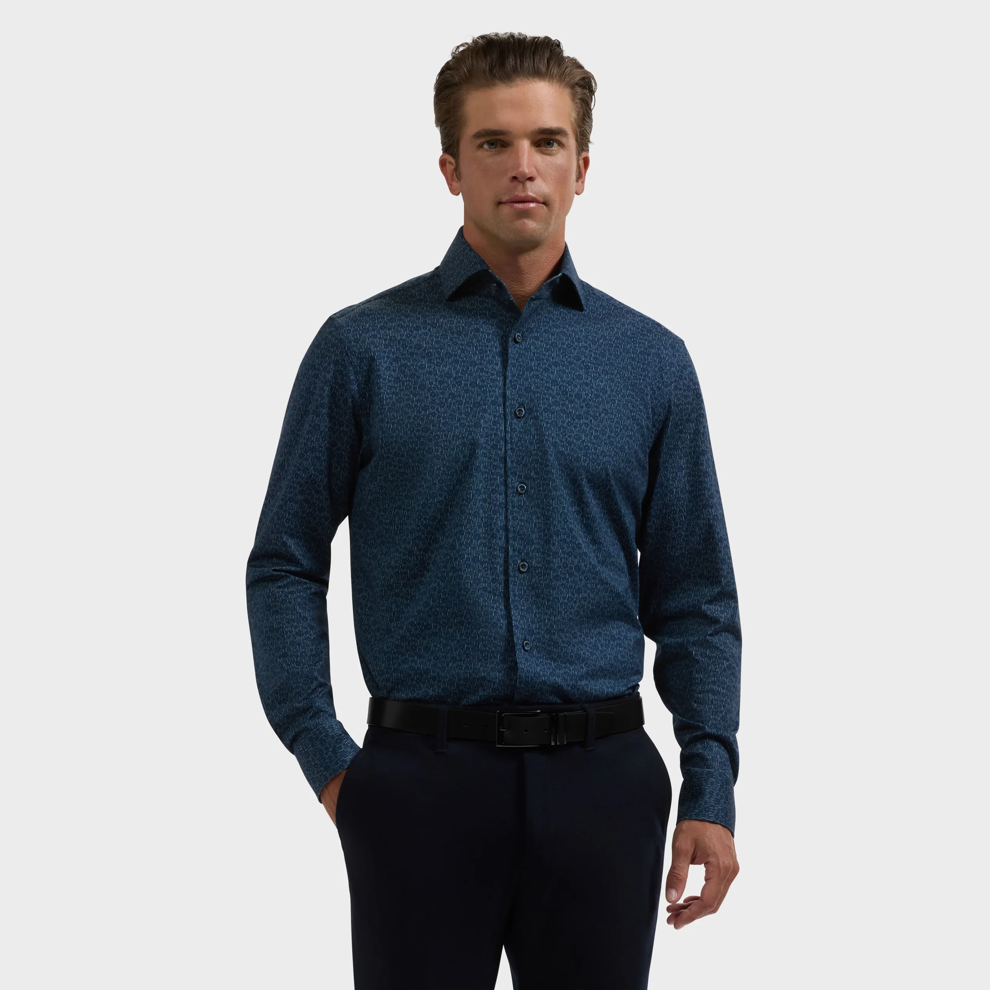 A man stands against a plain background wearing the Bugatchi James Floral Sport Shirt, a dark blue patterned design crafted from wrinkle-resistant performance fabric by Bugatchi, neatly tucked into black pants with a matching black belt for a sharp, polished look all day.