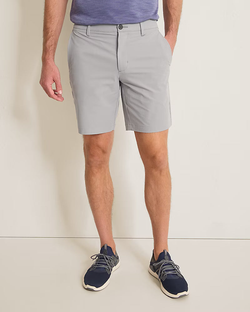 A man wearing Tommy Bahama Barbados Shorts Big & Tall in light gray, a blue shirt, and navy sneakers stands against a plain light-colored wall with his hands in his pockets.