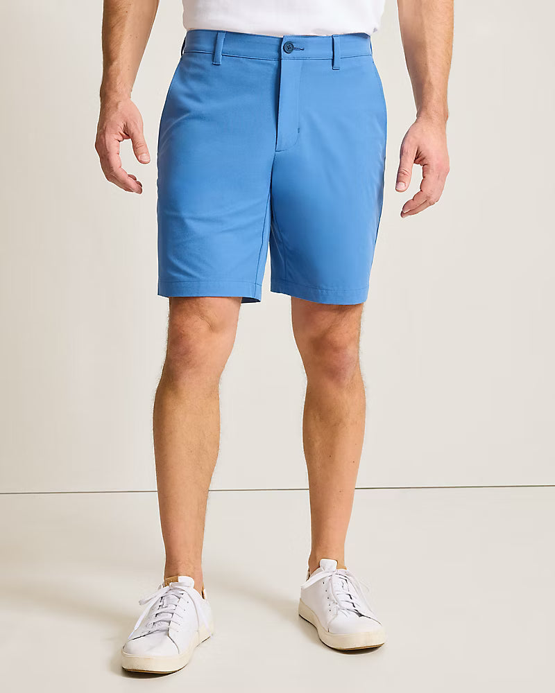 A man wearing bright blue Tommy Bahama Barbados Shorts Big & Tall, a tucked-in shirt, and white sneakers stands against a plain light background.