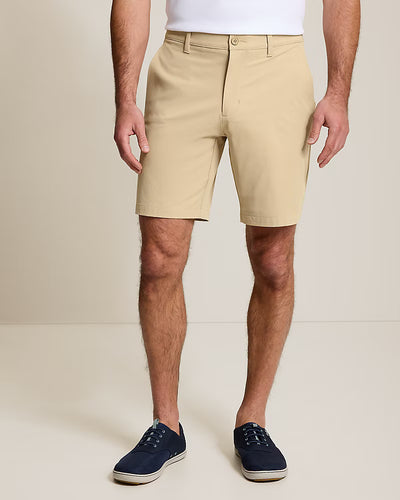 A person in beige knee-length Tommy Bahama Barbados Shorts Big & Tall, paired with a white shirt and navy blue sneakers, stands against a plain light background.