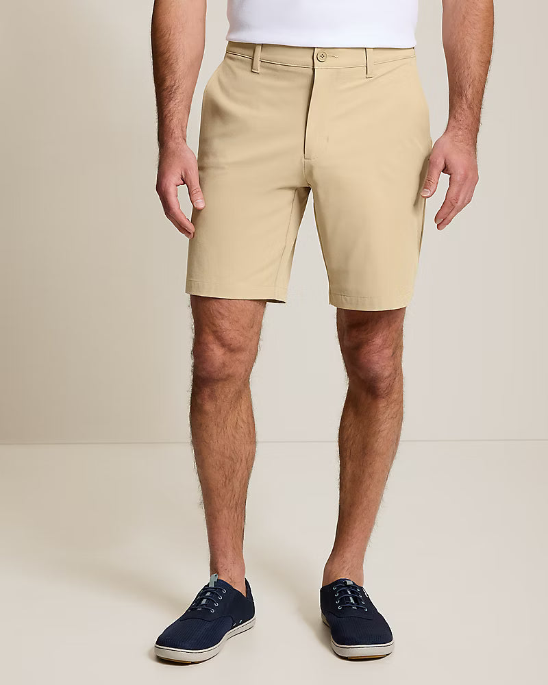 A person in beige knee-length Tommy Bahama Barbados Shorts Big & Tall, paired with a white shirt and navy blue sneakers, stands against a plain light background.