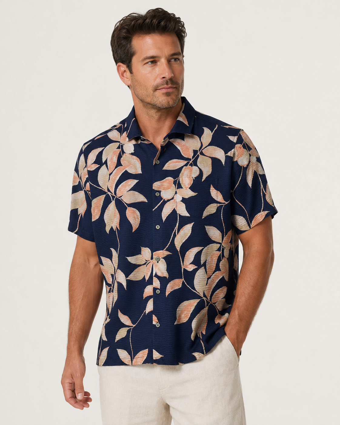 A man wearing the Tori Richard Textured Fresco De Cas Shirt 1956M0387—a navy blue, short-sleeve button-up crafted from cotton pucker lawn and featuring a beige and peach leaf pattern—paired with light-colored pants, stands against a plain background.