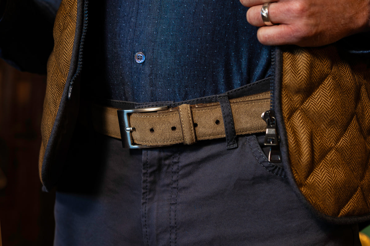 Close-up of a person wearing navy pants, a blue shirt, and the Martin Dingman Royal Suede Belt, with classic styling and one hand adjusting their brown textured vest.